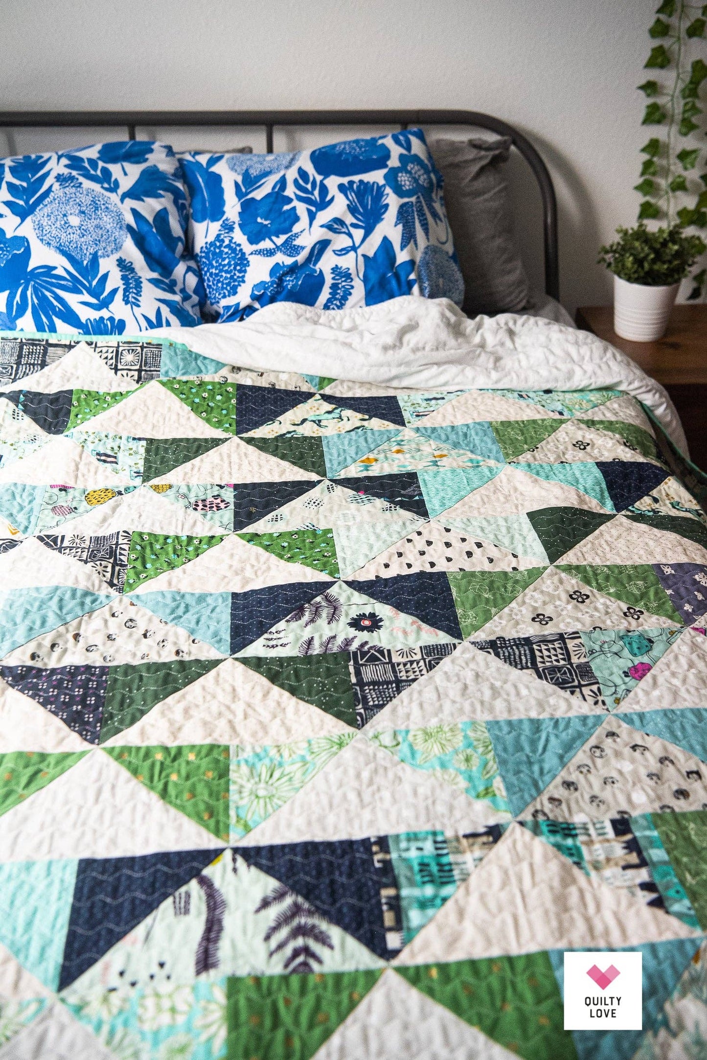 Quilt with geometric pattern on a bed with floral pillows and a plant in the background.