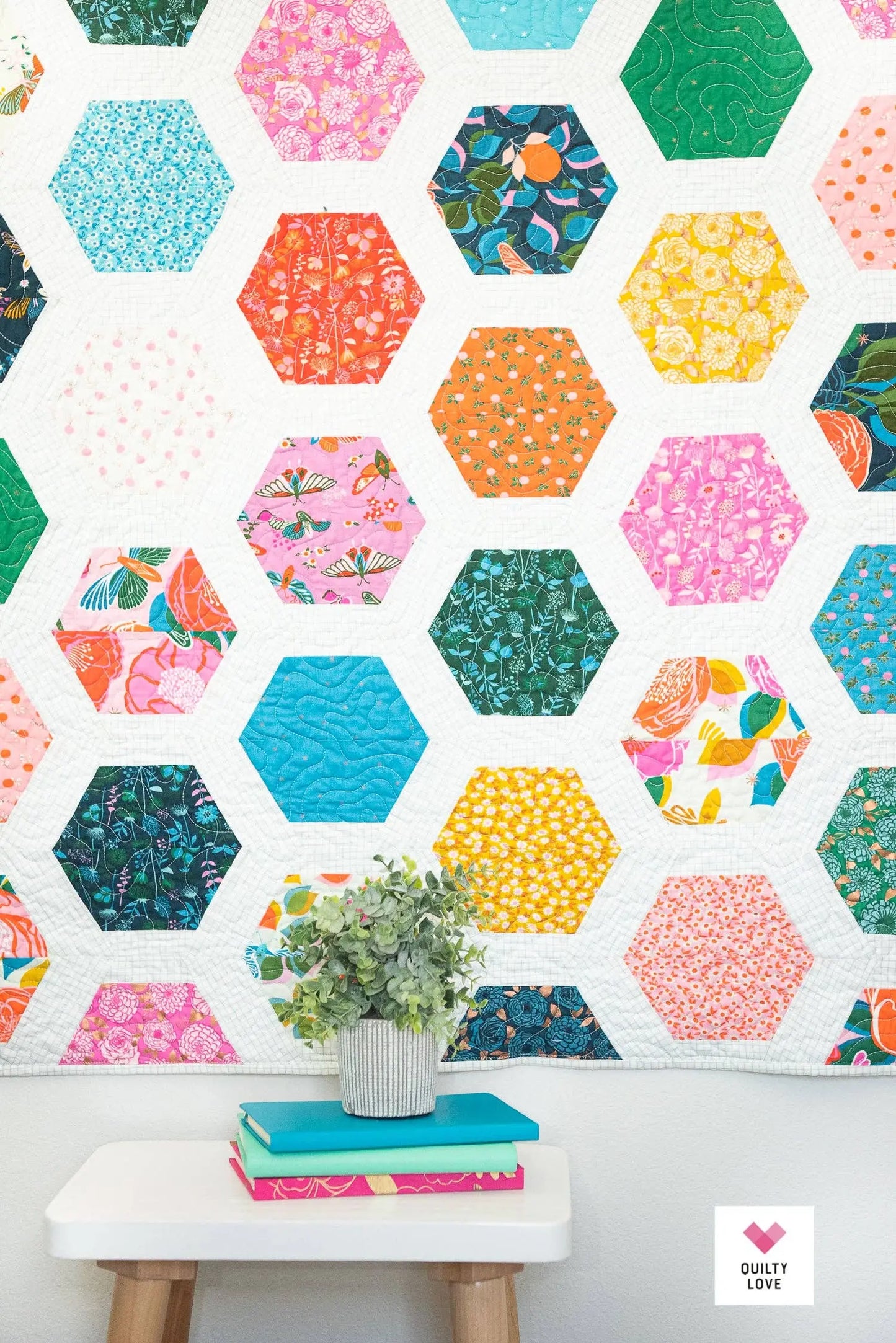 Hexagonal patterned quilt with colorful designs on a white wall, featuring a small table and books below.