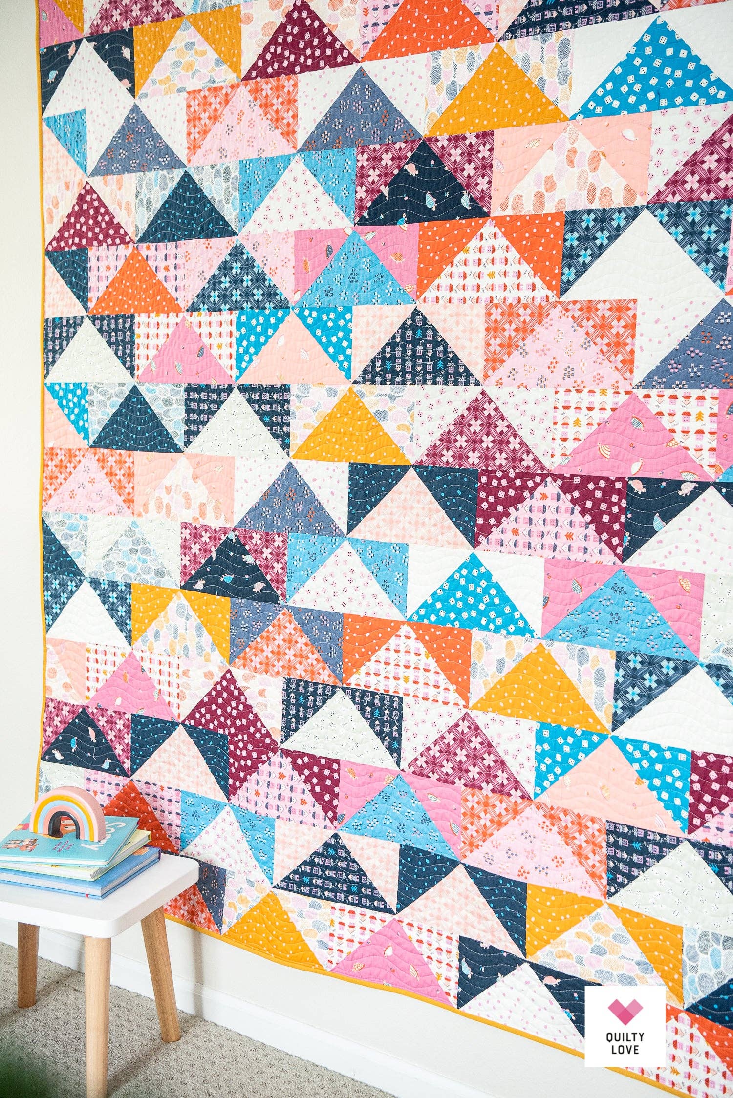 Colorful geometric triangle quilt on a wall with a small table and books in the foreground.