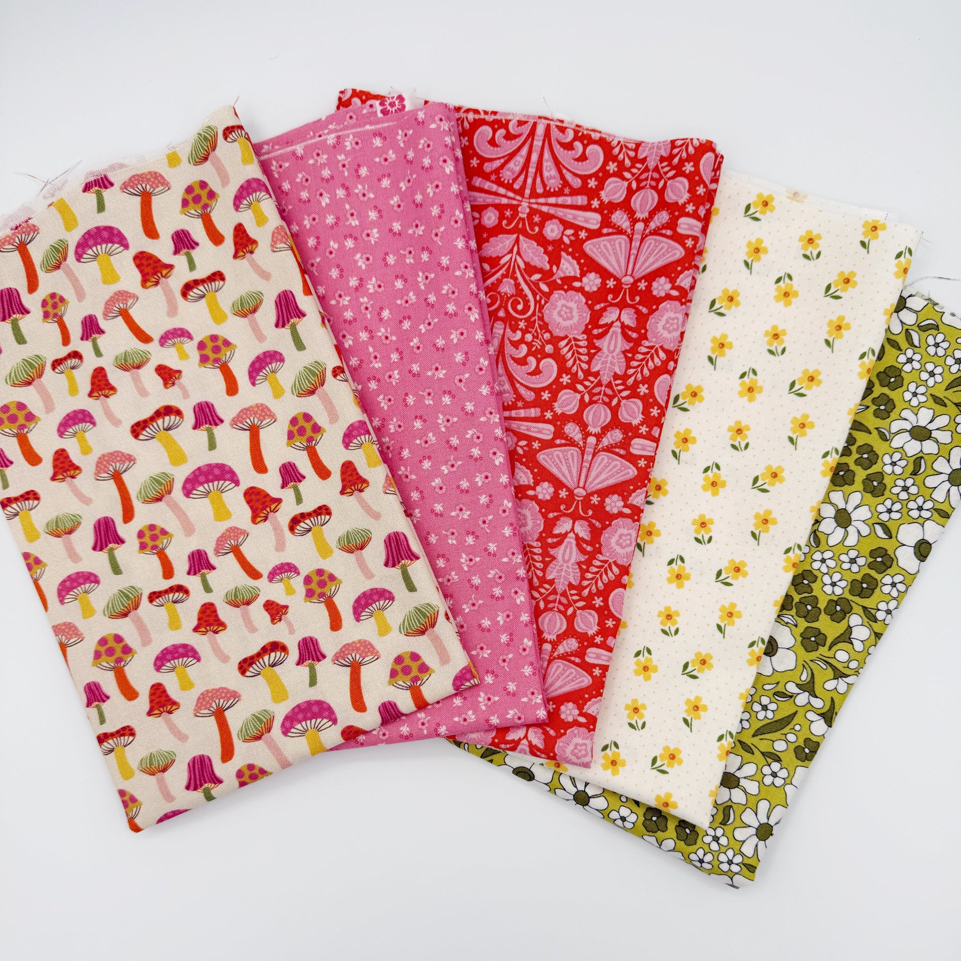 Fanned stack of coordinating quilting cotton fat quarters featuring cream mushroom print, pink ditsy floral, red folk art floral, cream fabric with yellow flowers, and olive green daisy floral