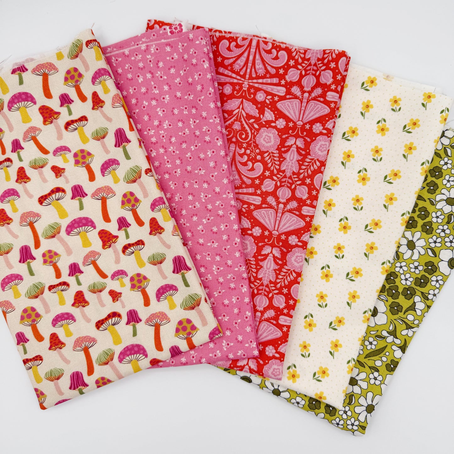Fanned stack of coordinating quilting cotton fat quarters featuring cream mushroom print, pink ditsy floral, red folk art floral, cream fabric with yellow flowers, and olive green daisy floral