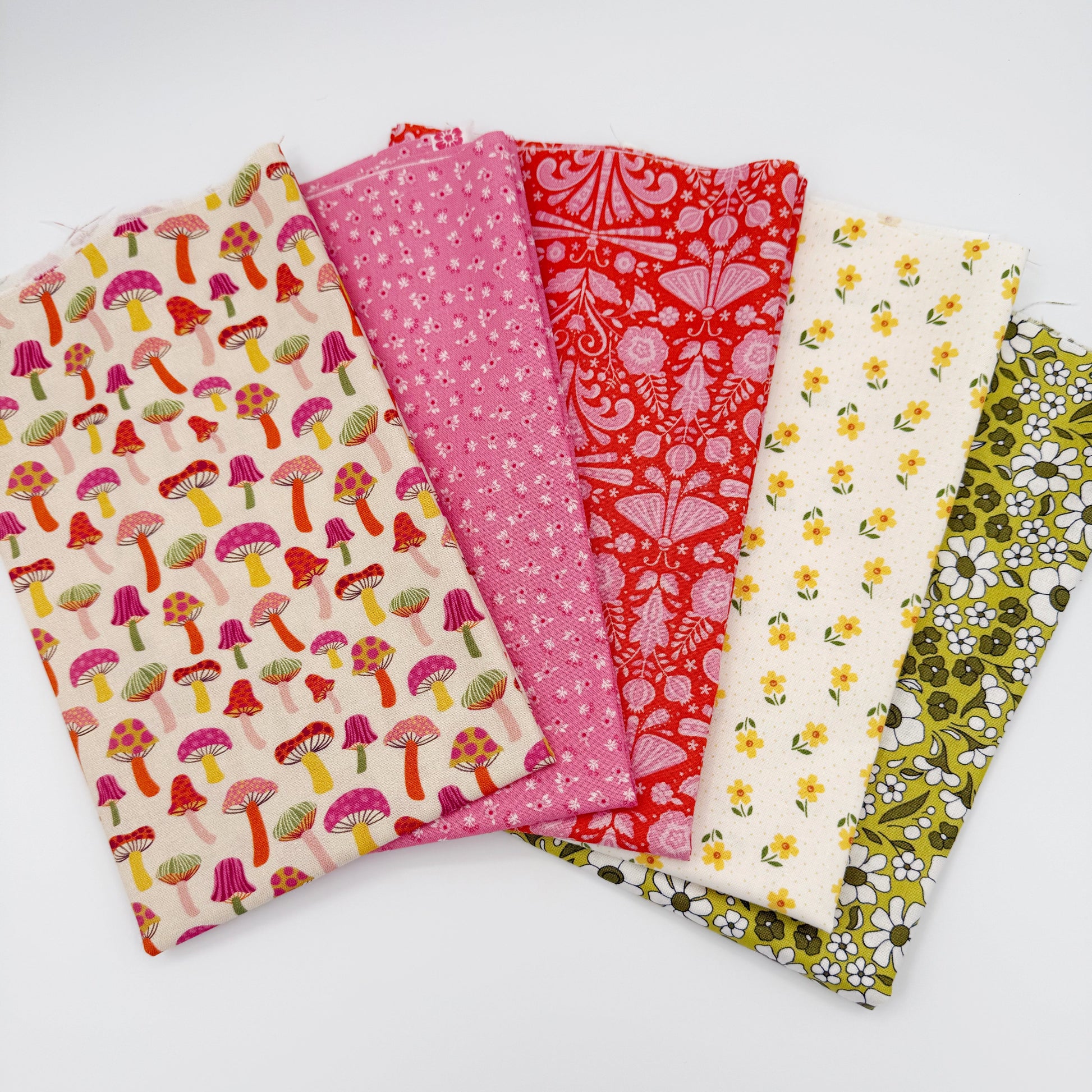Fanned stack of coordinating quilting cotton fat quarters featuring cream mushroom print, pink ditsy floral, red folk art floral, cream fabric with yellow flowers, and olive green daisy floral