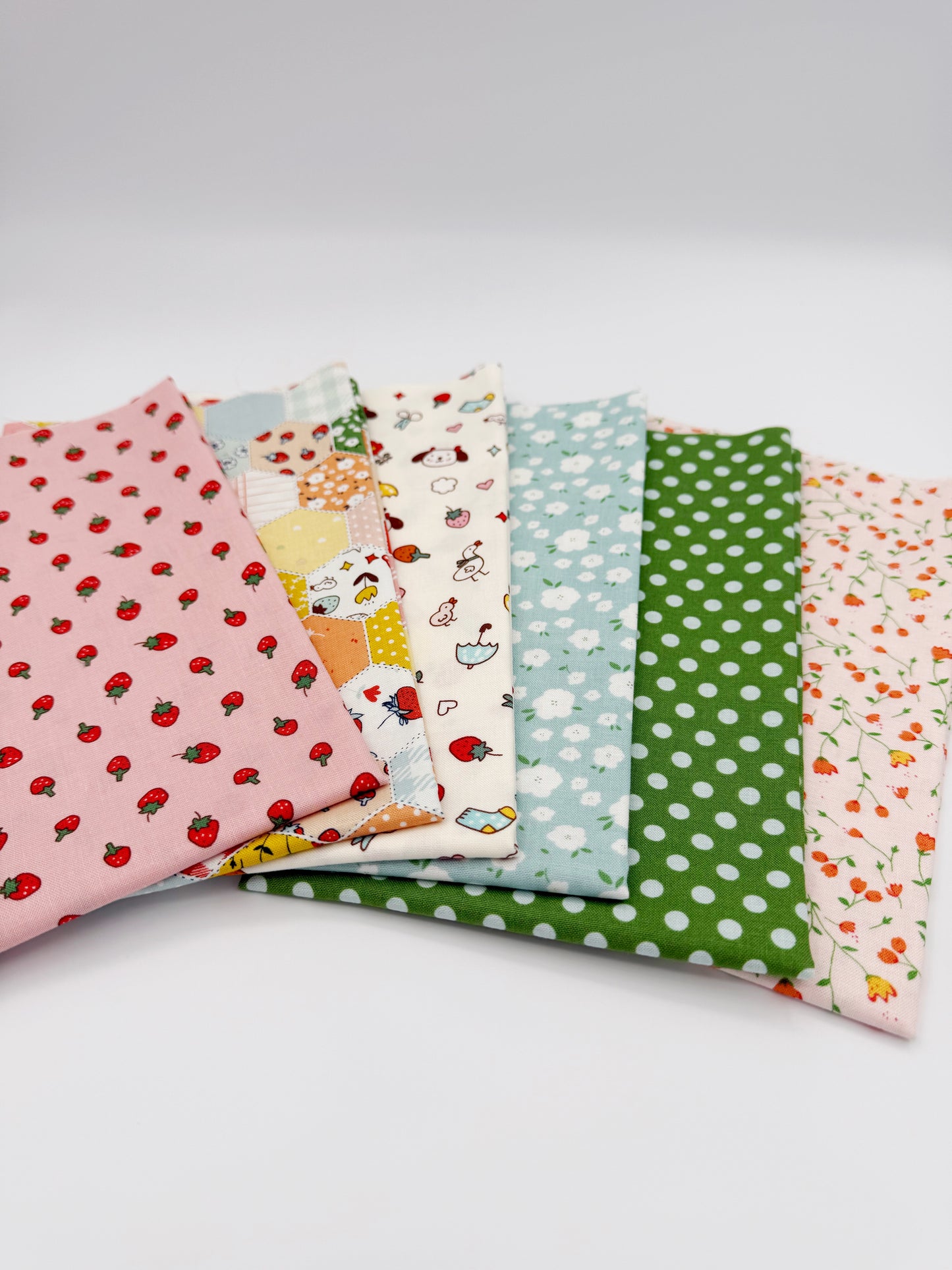 Fanned stack of coordinating quilting cotton fat quarters featuring pink strawberry print, patchwork hexagon print, cream fabric with dogs and strawberries, blue floral print, green polka dot, and pink floral vine print