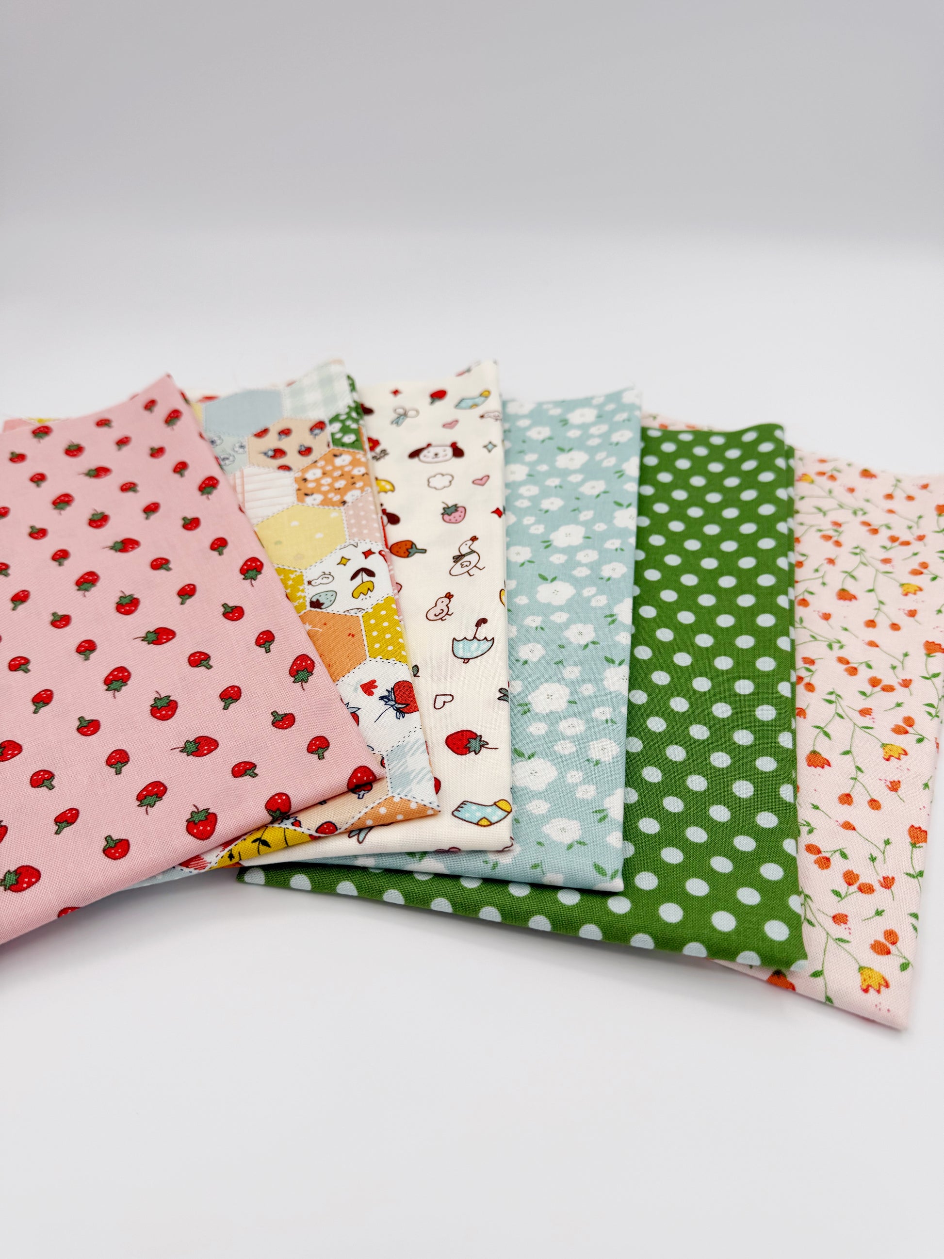 Fanned stack of coordinating quilting cotton fat quarters featuring pink strawberry print, patchwork hexagon print, cream fabric with dogs and strawberries, blue floral print, green polka dot, and pink floral vine print