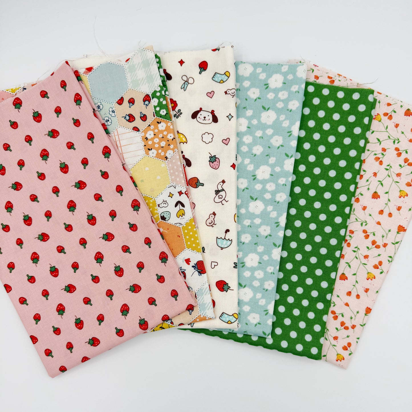 Fanned stack of coordinating quilting cotton fat quarters featuring pink strawberry print, patchwork hexagon print, cream fabric with dogs and strawberries, blue floral print, green polka dot, and pink floral vine print