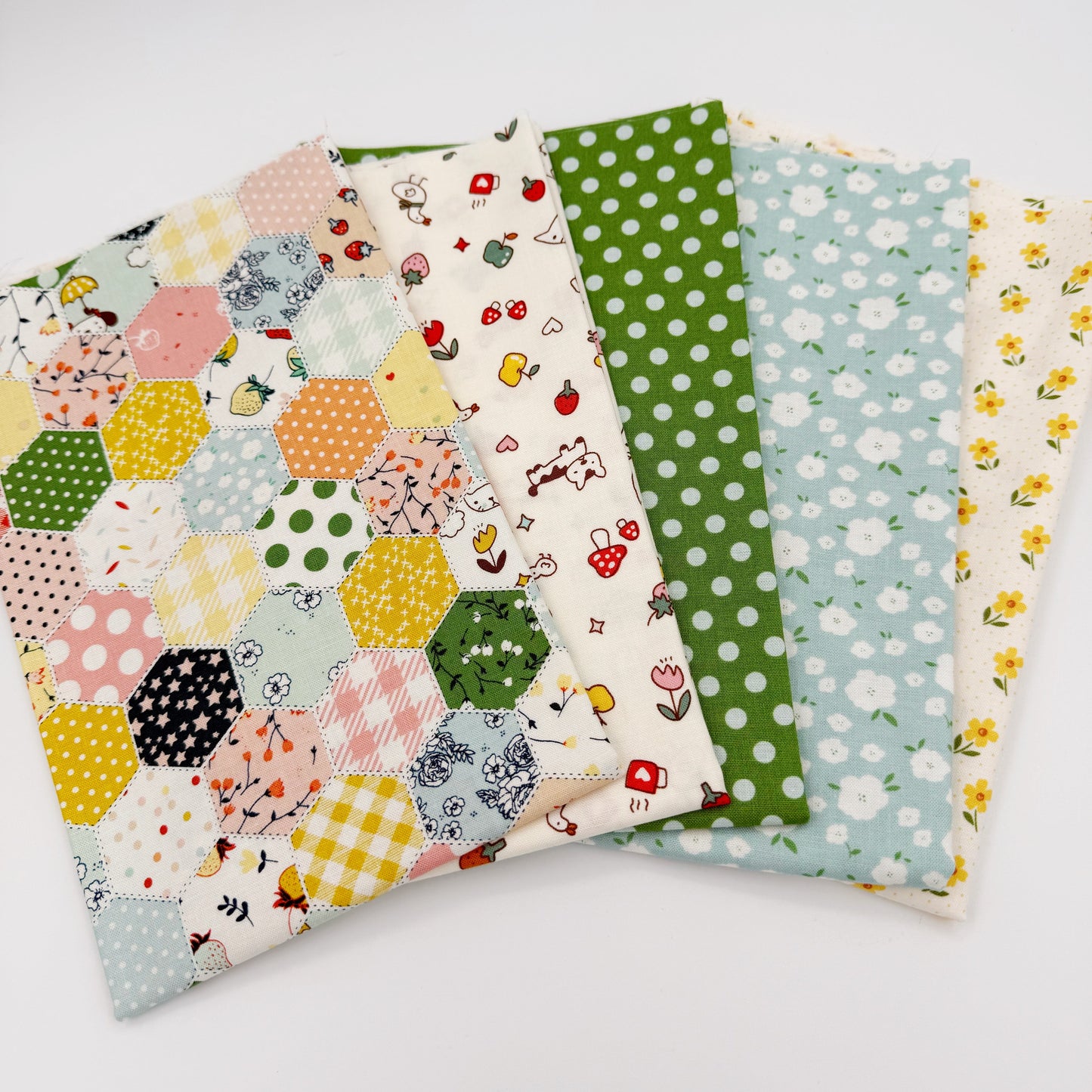 Fanned stack of coordinating quilting cotton fat quarters featuring multicolor patchwork hexagon print, cream fabric with strawberries and puppy, green polka dot, blue floral, and cream fabric with yellow flowers