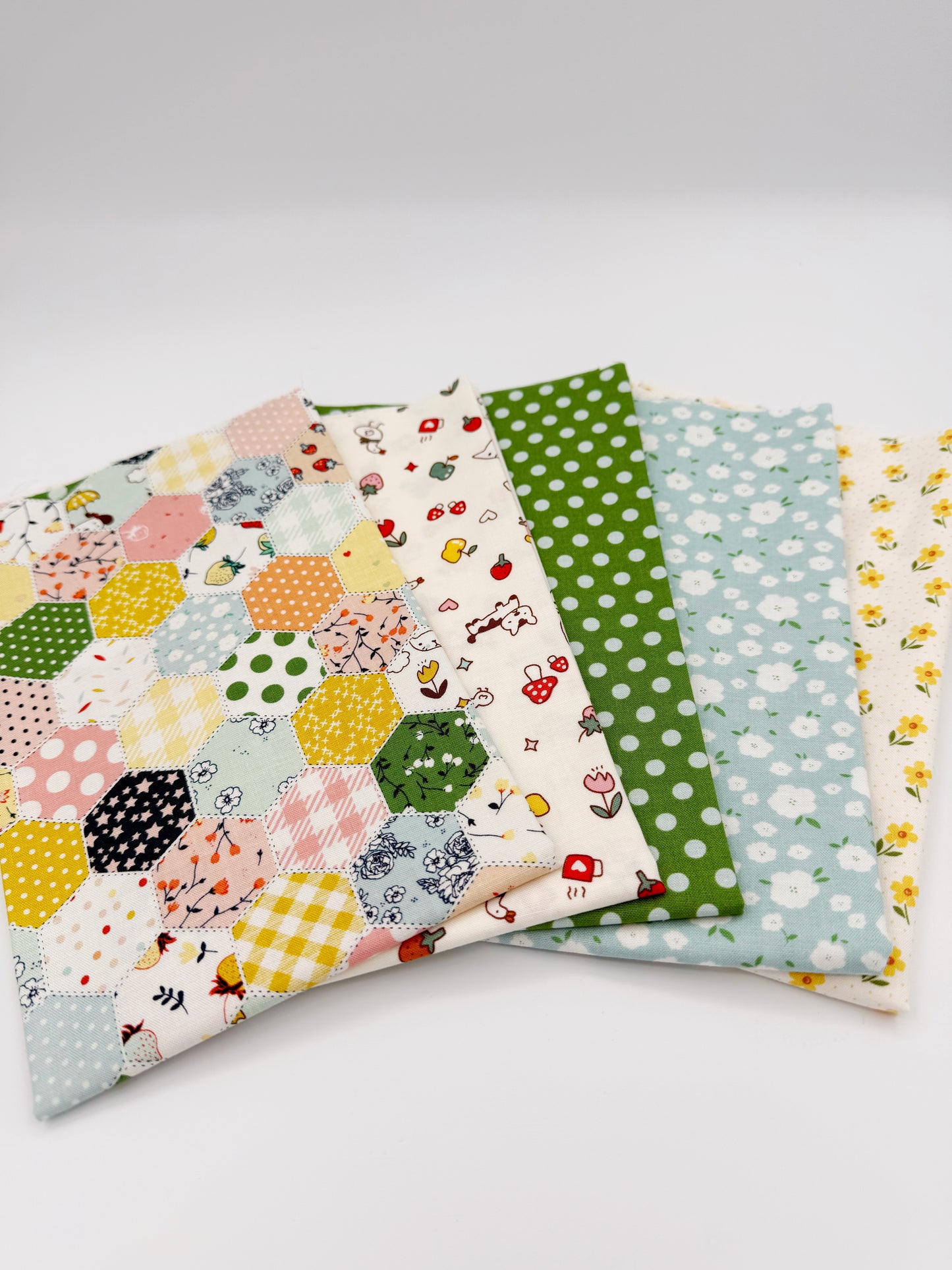 Fanned stack of coordinating quilting cotton fat quarters featuring multicolor patchwork hexagon print, cream fabric with strawberries and puppy, green polka dot, blue floral, and cream fabric with yellow flowers