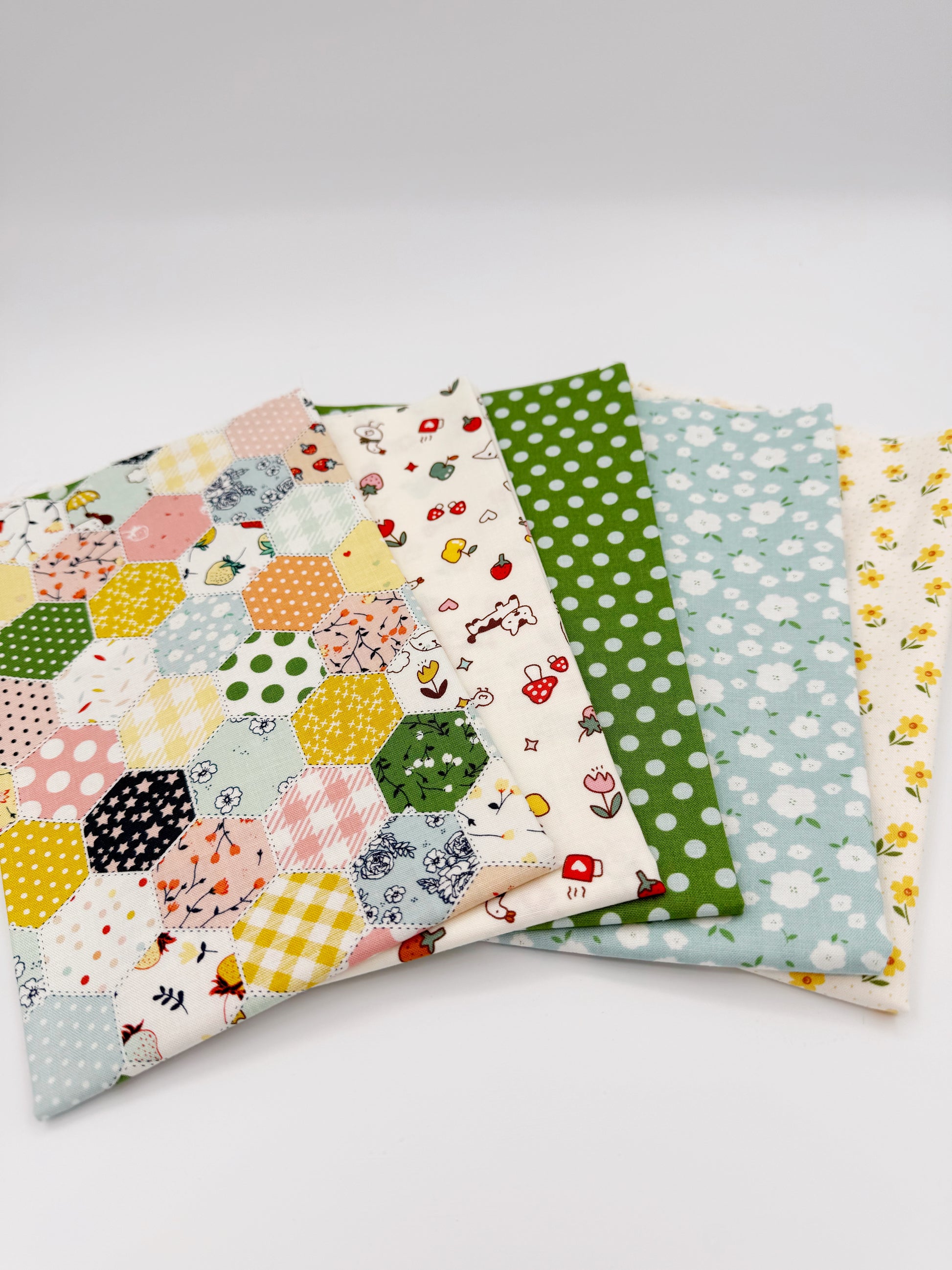 Fanned stack of coordinating quilting cotton fat quarters featuring multicolor patchwork hexagon print, cream fabric with strawberries and puppy, green polka dot, blue floral, and cream fabric with yellow flowers