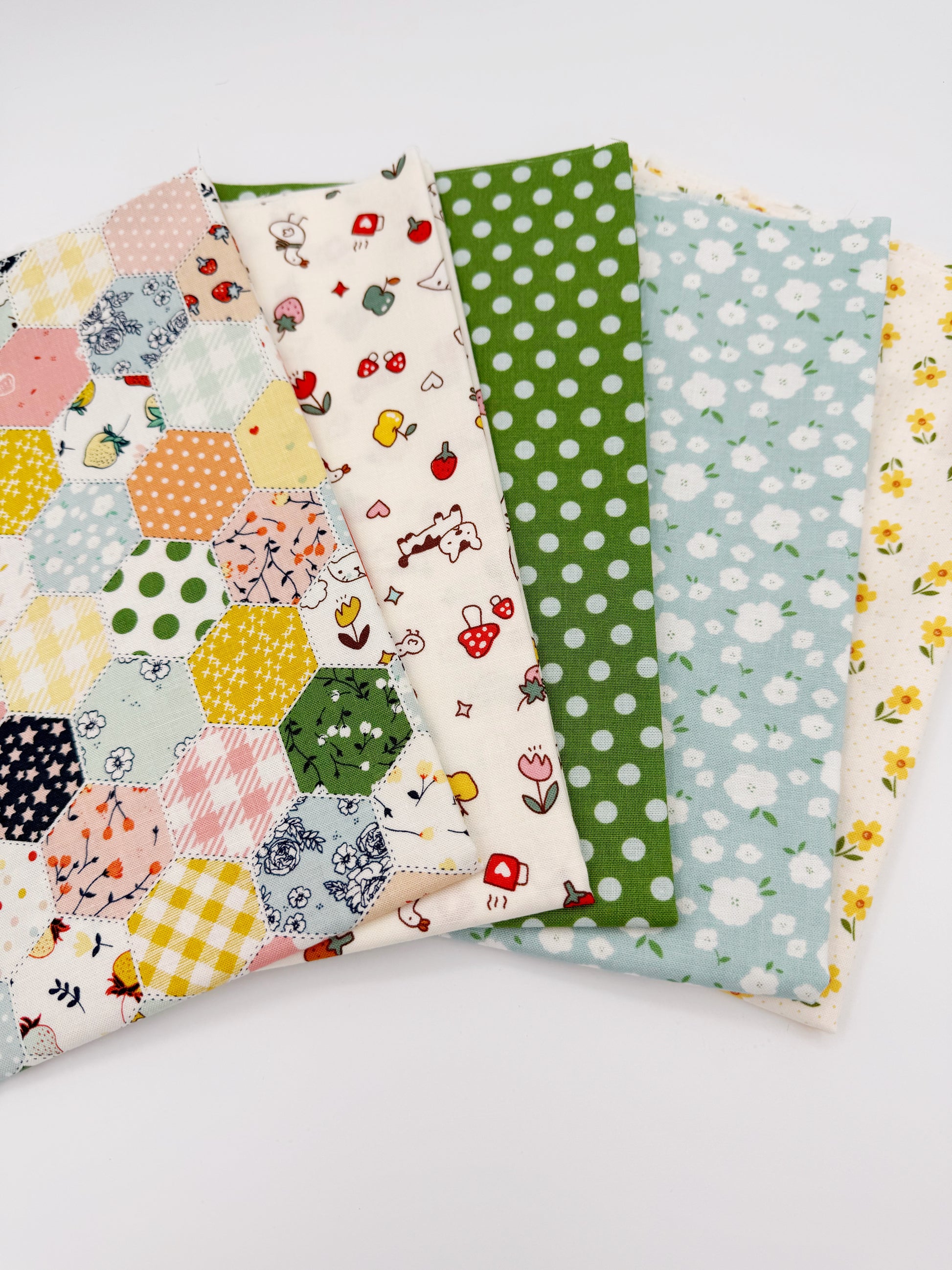 Fanned stack of coordinating quilting cotton fat quarters featuring multicolor patchwork hexagon print, cream fabric with strawberries and puppy, green polka dot, blue floral, and cream fabric with yellow flowers