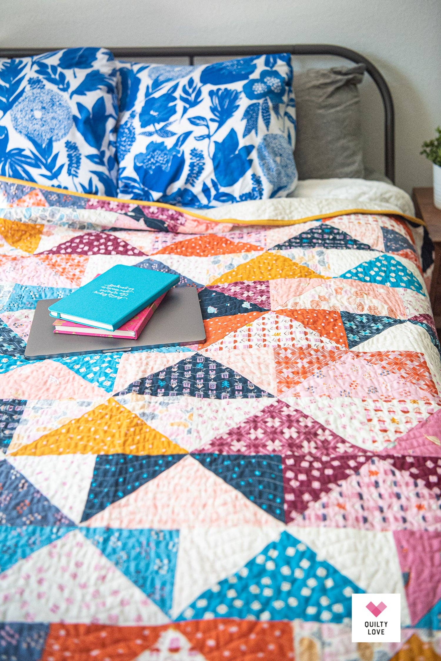 Colorful quilt with geometric pattern on a bed with floral pillows and books.